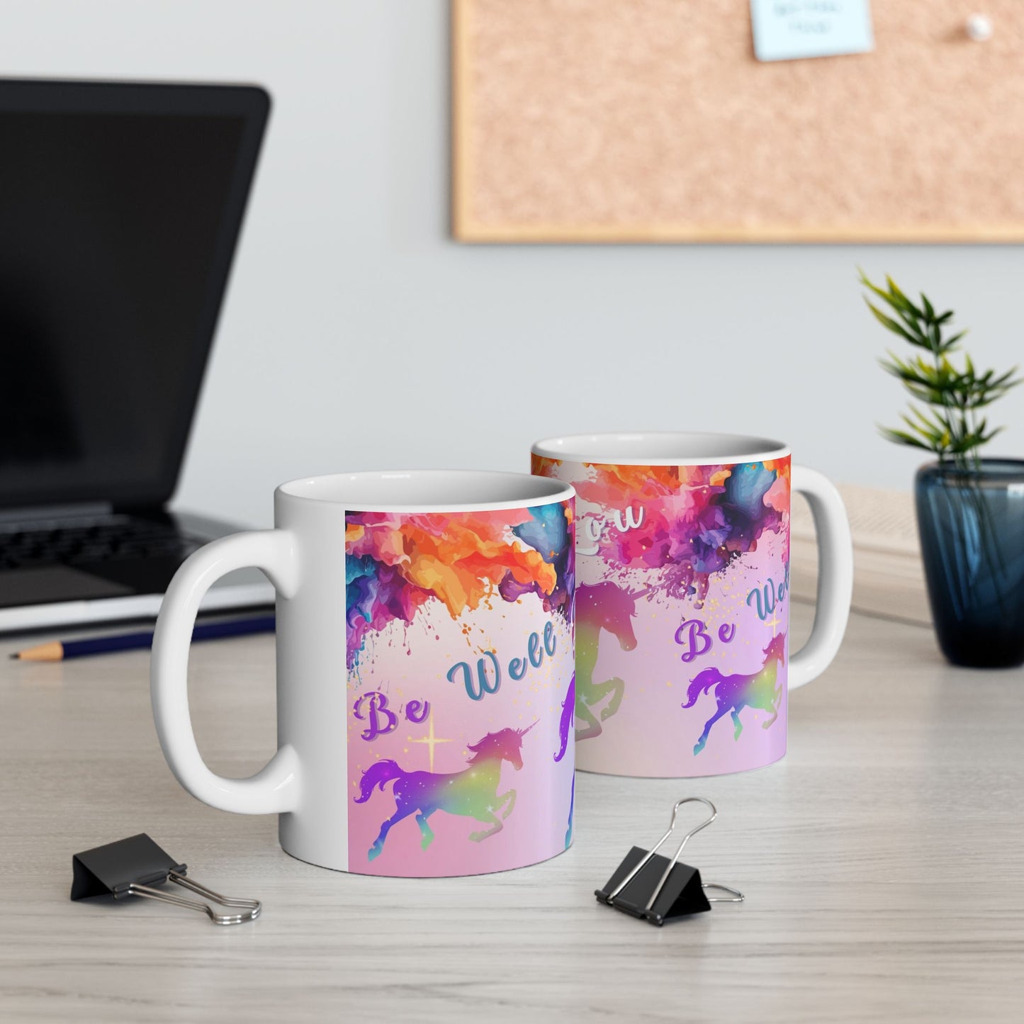 Two white ceramic mugs with colorful watercolor splashes and unicorn designs, displayed on a light wood desk with office props like a laptop, binder clips, and a small plant.