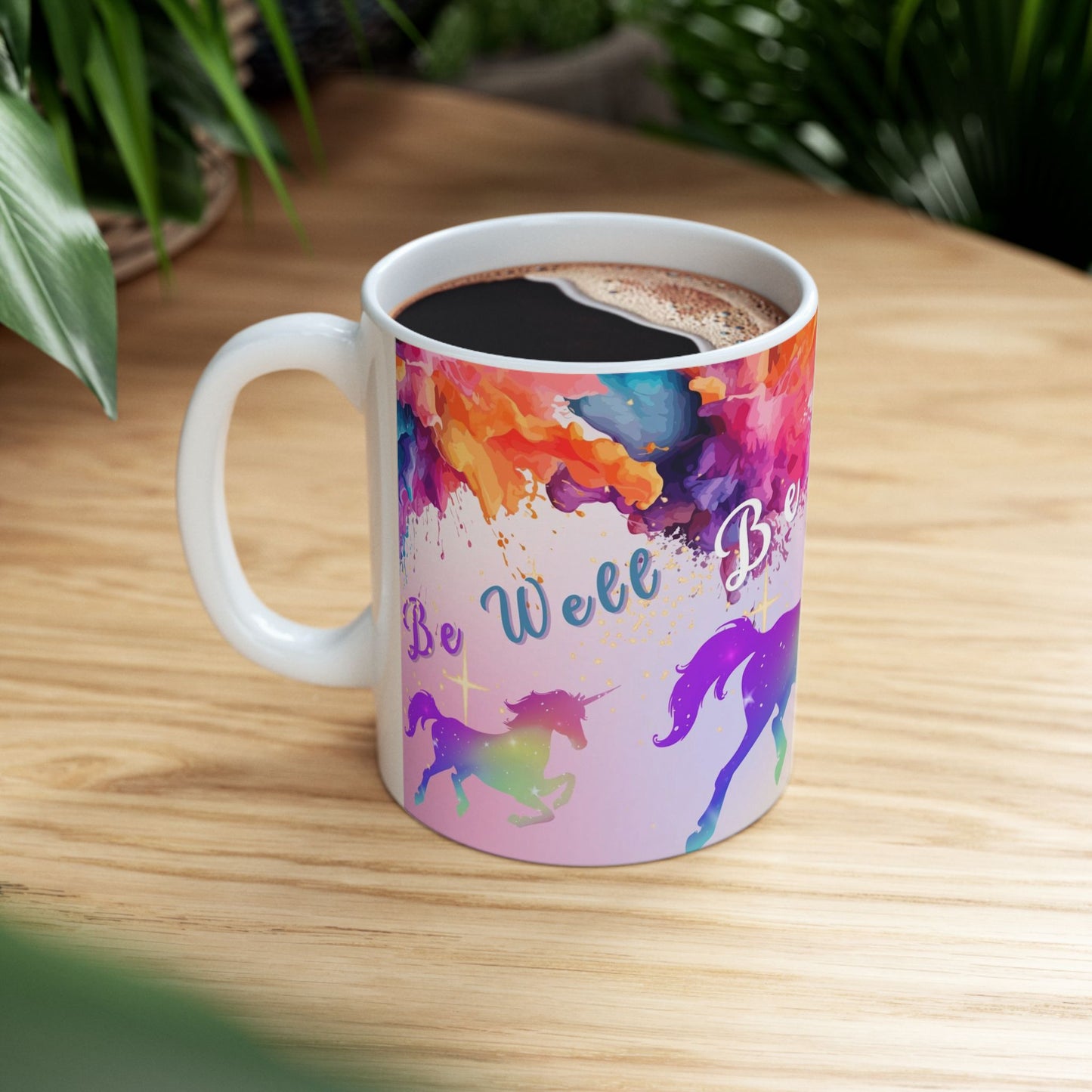 A white ceramic coffee mug filled with a dark beverage, featuring a colorful pastel watercolor design with two unicorns and the text 'Be Well Be Bold' on a light wood tabletop with blurred green plants in the background.
